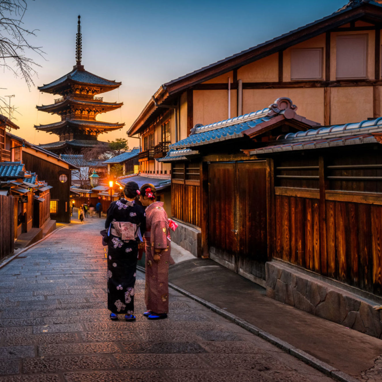 Two people in traditional Japanese attire walking along a street with a pagoda in the background.